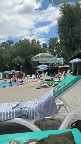The video opens with a view of a poolside area. In the foreground, a lounge chair is draped with a striped towel. The camera pans slightly, revealing more lounge chairs and umbrellas. Beyond the chairs, a swimming pool is visible with several people in and around it. A woman in a pink bikini is sitting on a lounge chair to the right. Further to the left, in the pool area, a group of people are gathered, and a person in an Iron Man costume is interacting with others. The environment is outdoors with trees and a cloudy sky.