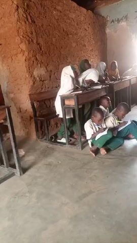 In a classroom, several children dressed in school uniforms are seated at wooden desks or on the floor. The classroom walls are roughly textured, appearing to be made of mud or a similar material. Some of the children wear head coverings, while others are bareheaded. All of them are writing on paper, seemingly engaged in a classroom activity. The room is lit by natural light coming through a window.