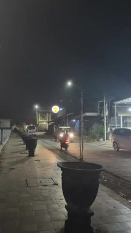 A vertical video captures a street scene at night. The foreground shows a tiled walkway lined with large, dark planters. Beyond the walkway, a street is visible with a few vehicles and buildings. A white van and a motorcycle with its headlight on are moving along the street. A streetlamp illuminates the scene. The sky is dark.