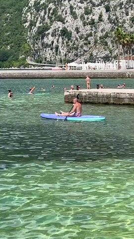 This video captures a sunny day at a coastal location, likely Risan, Montenegro. The primary subject is a man paddleboarding in clear, turquoise water. He is seated on the board, holding a paddle and appears to be casually moving through the water.

In the background, several other people are visible enjoying the water. Some are swimming, and others are near a stone jetty. One person is standing on the jetty, and a few others are in the water near it. A sailboat with a white sail is docked or anchored in the distance, adding to the nautical atmosphere.

The setting is picturesque, with lush green hills rising steeply behind the coastline. The buildings on the hillside suggest a populated area, possibly a town or village. The water is remarkably clear, allowing viewers to see the dappled light patterns on the seabed. The bright sunlight and clear water indicate warm, pleasant weather, suitable for water activities. The overall mood is relaxed and leisurely, typical of a vacation or a summer day spent by the sea. There is no discernible text visible in the video.