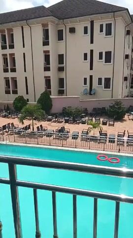 The camera, positioned behind a metal railing, offers a view of a rectangular swimming pool filled with bright blue water. Two red life preservers float in the water. The pool is surrounded by a tiled patio dotted with black tables and chairs, as well as small palm trees and bushes. Behind the patio is a tall, light-colored building with multiple windows and several air conditioning units mounted on its exterior walls. The scene appears to be taken outdoors on a sunny day.