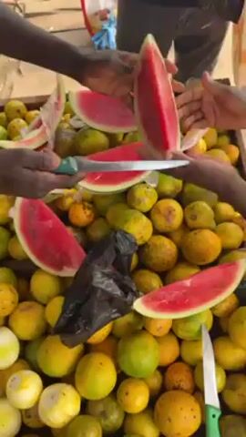 A cart displays a collection of green and yellow mangoes, with watermelon slices arranged on top. A person is shown cutting another slice of watermelon with a knife. Another knife is lying amongst the mangoes. The scene is outdoors.