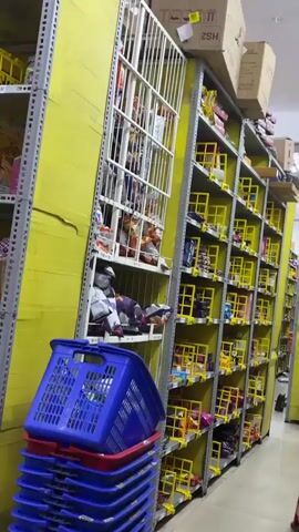The video displays an interior view of a retail store aisle. Tall, metallic shelving units with bright yellow back panels line the right side of the frame, extending into the background. These shelves are densely packed with numerous small, individually packaged products, many of which are displayed within yellow grid-like plastic compartments or dividers. Another set of shelves, less distinctly visible and holding various items, is present on the left side. In the bottom left foreground, a stack of blue and red plastic shopping baskets is partially visible. The camera slowly pans across the aisle, showcasing the extensive product display. No people are visible in the scene.