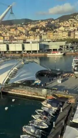 The video shows a harbor scene. In the foreground, several small white motorboats are docked at a wooden pier. Behind the pier, there is a large white structure resembling a canopy or tent, supported by a series of white poles. People are visible underneath this structure, but their actions are not clearly discernible due to the distance.  In the background, there is a city built on a hillside, with buildings of various colors and styles. The sky is blue with a few scattered clouds, suggesting a sunny day. The camera appears to be positioned at an elevated viewpoint, possibly from a building or a tall structure overlooking the harbor.