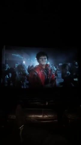 A person is sitting in a movie theater with their legs stretched out and watching Michael Jackson's Thriller music video on the big screen. The screen is showing Jackson dancing with zombies on a street at night. The theater appears dark, with rows of empty seats in front of the viewer. Subtitles in Chinese are visible at the bottom of the screen.