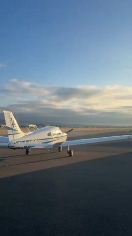 A small white airplane with blue accents is parked on an asphalt tarmac. The airplane has a single propeller at the front. The sky is clear with some scattered clouds, and the sun appears to be low in the sky, casting long shadows. In the background, there are buildings and hills. The video pans slightly to show different views of the airplane and its surroundings.