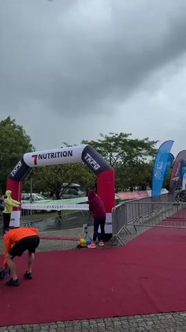 In a medium shot, a runner in triathlon gear crosses the finish line under an arch that reads “7 NUTRITION” and “TKFG” on a rainy day. A red carpet spans the finish line. Several people, including photographers, are present at the finish line. Some individuals are wearing rain gear. The backdrop features trees, parked cars, and overcast skies. The camera is positioned low, looking upwards towards the finish line.