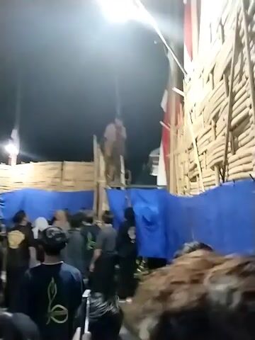 The video shows an outdoor night event in Jenggrik, Indonesia, featuring a cultural performance. The scene is illuminated by bright artificial lights, with temporary structures made of bamboo and blue tarps forming a backdrop. Indonesian flags are visible on these structures.

Initially, a person in traditional attire is seen being lowered from a height by a rope or harness, descending onto a dirt ground. A crowd of people, mostly young men dressed in casual clothing, watches the descent. Once on the ground, the person in traditional costume begins to move rhythmically, joining the crowd. Many individuals in the crowd also move and sway, indicating a lively atmosphere. The event appears to be a gathering or celebration, possibly a traditional dance or ritual.