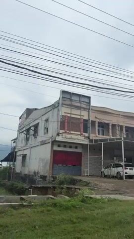 This media captures a street view in Lampeuneurut Blang, Indonesia, under an overcast sky, suggesting it's daytime. The scene is lined with buildings, some appearing to be commercial establishments.

On the left, a building with a pink and green facade features an air conditioning unit and windows. Beneath it, a white car is parked under a covered area. Further down the street, a black SUV is parked in front of a building with a sign that reads "PONDOK LAUNDRY".

The middle section of the video shows a building with a large sign for "RAHMAT JAYA PAINT & RESTORATION" with a Vespa logo. Underneath this, several motorcycles are parked, and two men are visible, one standing and another near some stacked plastic chairs and water containers.

Towards the right, another building has signs, one partially obscured, and a utility pole. The scene ends with a glimpse of lush green trees and foliage, indicating a mix of urban and natural elements in the environment. The overall impression is a quiet, ordinary day on a street with various businesses and parked vehicles.