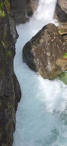 The video captures an overhead view looking down into a narrow, rocky gorge where a powerful torrent of water flows. Large, dark, wet boulders dominate the frame, with the turbulent, white water of a waterfall or rapid crashing between them. On the left, water cascades over a rock face, creating significant spray. On the right, a large, rounded boulder diverts a strong flow of water. The water is foamy and appears light blue in calmer sections, while white where it's turbulent. The surrounding rocks are rugged and dark, with hints of green moss and small plants growing in crevices and on ledges, particularly at the bottom edge of the frame. The scene is illuminated by daylight, suggesting a clear or partly cloudy day. There are no people or artificial objects visible, emphasizing the raw power and beauty of nature. The location is a natural landscape, consistent with Sahún, Spain.