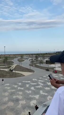 The video begins with a close-up of a blue toy airplane mid-air against a cloudy sky. The camera then pans down to show a young person wearing a white t-shirt and a dark baseball cap looking up at the sky, arm extended as if throwing the airplane. The scene is an outdoor public space with winding paved pathways, scattered trees, lampposts, and benches. In the background, a body of water, likely the sea, is visible. Several other people are walking in the paved area below. The perspective shifts to show the back of the young person's head, with another person to their right holding a smartphone. The camera continues to capture the park area with more people walking around. The last shot briefly shows the young person's head and shoulders from the side, looking down.