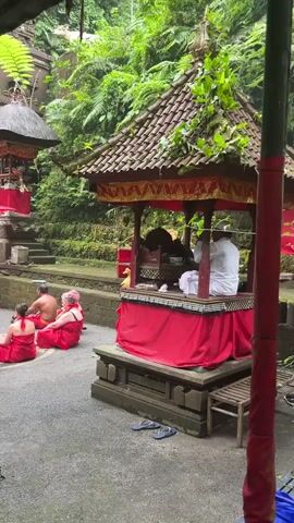 A group of people wearing red sarongs are sitting on the ground facing a structure with a red and gold roof. A person in a white outfit is seated inside the structure. The environment appears to be outdoors with lush green foliage.
