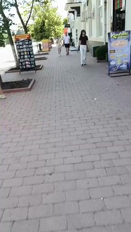 The video shows a person walking on a brick sidewalk lined with trees and a building on the right. A currency exchange board with various currency rates is visible on the left. Several people are walking away from the camera on the sidewalk. The environment appears to be an urban street on a sunny day.