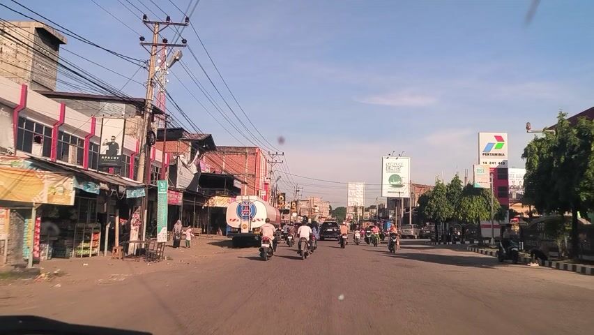 The image presents an inverted, bright daytime view of a bustling street in Meukokuthang, Indonesia, likely captured from a moving vehicle. The sky is clear and blue, indicating sunny weather.

The scene is dominated by traffic and commercial activity. Numerous people are visible riding motorcycles, which are a primary mode of transportation on the road. A large white tanker truck with a blue and red logo is also prominent on the right side of the street, suggesting transport or delivery services.

Buildings line both sides of the road, appearing to be a mix of residential and commercial structures, many with storefronts at ground level. Utility poles and a dense network of overhead wires crisscross the view.

Notable visible text includes:
*   An inverted billboard on the left displaying "PERTAMINA" and "ISI," referring to the Indonesian state-owned oil and gas company.
*   Another green billboard further down the left side, showing partially legible text that includes "MAS" or "EMAS."
*   The logo on the tanker truck is a distinctive blue and red emblem, but specific text is not clear.

The image captures a typical lively street scene in an Indonesian town or city during daylight hours.