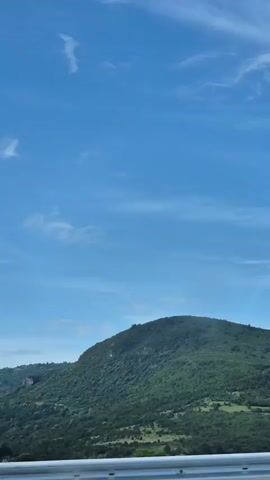 The video shows a green, treed hill against a blue sky with wispy clouds. A white railing is visible at the bottom of the frame. The camera pans to the right, showing more of the hill and sky.