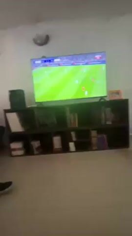 The video shows a person watching a soccer game on television. The TV is placed on top of a brown bookshelf filled with books. The camera zooms in to show the screen. A soccer player wearing a green uniform is seen kicking the ball on the field. The game is between Real Madrid and Benfica. The room appears to be a living room with a neutral colored floor and white walls. A black speaker sits next to the TV on top of the bookshelf.
