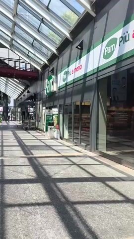 The video shows a view inside a covered walkway or mall. A glass roof allows sunlight to stream in, creating distinct shadow patterns on the tiled floor. On the right side of the walkway, there are storefronts, including one with a green and white "Pam" logo and the text "Pam Più a meno". Further down, another "Pam" sign is visible. To the left, there's a cafe area with tables and chairs, and a sign that reads "La Galleria". Above the cafe, a large banner advertises "Fitness Formula" with images of a woman and a man, and text that says "BEN essere per tutti TI ASPETTIAMO AL 1° PIANO". In the distance, there are a few people walking, shopping carts, and what appear to be entrances or exits. The overall atmosphere seems to be that of a commercial area on a sunny day.