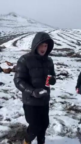 In a snowy landscape, a man stands wearing a black hooded puffer jacket, black pants, and brown boots. He has gloves on, holding a snowball in one hand and a Coca-Cola can in the other. In the background, a snow-covered hill can be seen with lines of trees. To the left, there are logs and some scattered debris on the ground. The man waves his hand to the camera while holding the coke can. The weather appears to be overcast.
