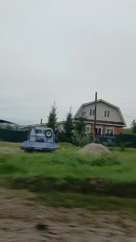 The video captures a passing view of a rural or suburban area, likely from a moving vehicle. The scene is characterized by overcast skies and lush green vegetation. Several houses and fences are visible, along with trees of varying sizes. A white van with a blue tarp-covered cargo area is parked on the right side of the frame. Another vehicle, possibly a flatbed truck, is also present. The road appears to be unpaved, with some puddles visible. The overall impression is that of a quiet, possibly damp, day in a residential area.