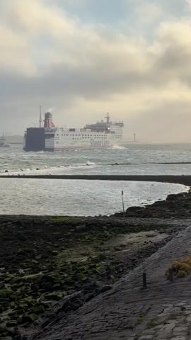 A large Stena Line ferry is seen sailing through choppy, grey water under a cloudy sky. Waves are crashing against a rocky shoreline in the foreground. A red buoy is visible in the water, marking a channel. In the distance, other ships and port infrastructure, including a lighthouse and cranes, can be seen. The scene suggests a busy maritime environment experiencing moderate weather conditions.