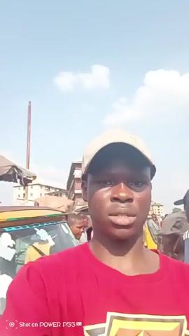 The video shows a young man standing in what appears to be a busy outdoor marketplace. He's wearing a red T-shirt, a tan baseball cap, and is looking directly at the camera. Behind him are other people walking around, and yellow auto-rickshaws, also known as tuk-tuks, are parked. Buildings and various stalls are visible in the background. The sky is blue with some white clouds. The video seems to be shot with a mobile phone, as indicated by the watermark at the bottom. The man raises his hand slightly as if acknowledging someone. The lighting is bright and the video appears to be taken during the day.