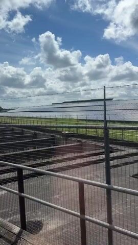 The video captures a moving perspective, likely from a vehicle, showing a landscape on a bright, sunny day with a blue sky and scattered white clouds. In the foreground, there are metal fences and structures that appear to be part of an enclosure or pens. Beyond these, a large field with solar panels is visible, stretching across the horizon. Further into the distance, there are trees and a small body of water. The camera pans and moves, offering glimpses of lush green vegetation and the changing scenery. The overall impression is one of a rural or industrial setting with elements of renewable energy infrastructure.