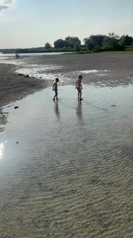The video shows a wide shot of a shallow body of water at what appears to be a lake or riverbank. Two children, a boy and a girl, are playing in the water. The boy is wearing blue swim trunks and is crouched down, seemingly looking at something in the water. The girl is wearing a pink swimsuit and is standing upright, facing away from the camera. The water is shallow, with ripples on the surface, and the bottom is sandy with some pebbles. In the background, there is a dry, muddy bank with patches of vegetation and trees lining the distant shore. The sky is blue with some scattered clouds. The sun is shining, casting reflections on the water. The video pans slightly to the right, revealing a wooden dock in the foreground.