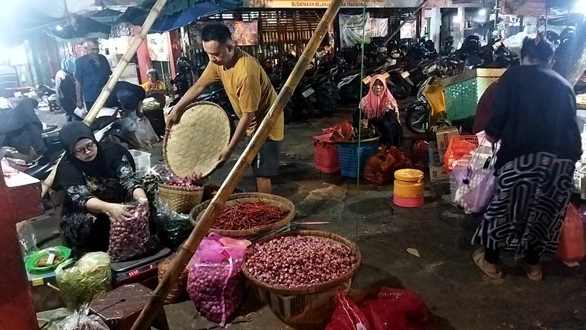 A bustling market scene unfolds at night. In the foreground, a man in a yellow t-shirt stands holding a large, flat, woven circular object. He is positioned between piles of red chili peppers and baskets overflowing with small red onions. To his left, a woman in a black hijab and glasses is seated, weighing produce on a scale, with bags of onions and green vegetables at her side. Behind her, other market-goers and stalls are visible. The background is filled with parked motorcycles, suggesting a busy commercial area. The lighting is dim, characteristic of an evening market. The overall atmosphere is one of active commerce and community gathering.