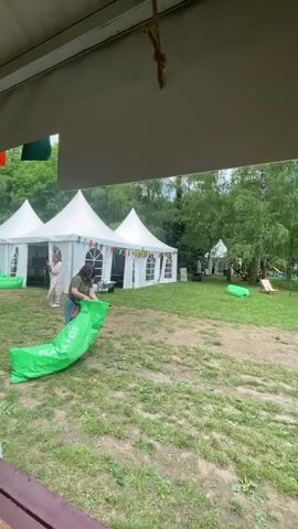 An outdoor scene depicts a grassy field with numerous green trees in the background. Three white peaked tents, adorned with colorful pennant banners, are visible on the left side of the frame. In the foreground, on the left, a person wearing a dark green t-shirt and light-colored pants is bending over a large, bright green inflatable object, appearing to handle it. Another person in light-colored clothing walks from left to right in front of the tents. Scattered across the grassy area are several wooden-framed lounge chairs with light fabric, along with additional bright green and one reddish-pink inflatable objects. The camera slowly pans to the right from the first person to reveal more of the outdoor setup. Distant voices and music are audible.