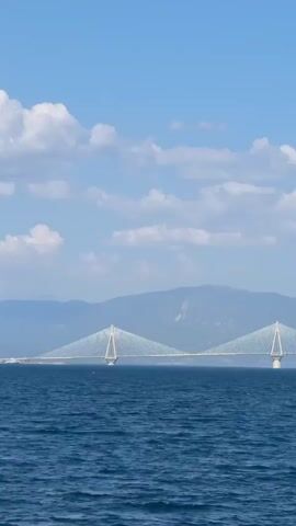 The video shows a cable-stayed bridge spanning a large body of water. The bridge has two prominent towers and a long deck supported by cables. The water is a deep blue, with gentle waves. In the background, there are mountains and a bright blue sky with scattered white clouds. The lighting suggests it is daytime. No people or vehicles are visible on the bridge or in the water.