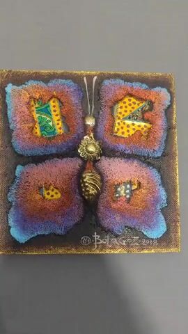 A close-up, slightly angled shot shows a decorative butterfly artwork mounted on a gray surface. The butterfly has large, four-part wings with ragged edges and a textured, fluffy appearance in shades of purple, pink, and blue, with touches of orange and yellow. Each wing section has an opening revealing fabric with patterns of yellow polka dots and green paisley. The butterfly's body is a central vertical element with a beaded antenna, a decorative metallic flower-like piece, and a ribbed, metallic lower body. The artwork is signed "@Botagoz 2017" in the lower center. The background is a plain, flat gray surface.