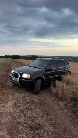 This video shows a black Suzuki Grand Vitara parked on a dirt road in a field of dry wheat. The vehicle has a chrome bull bar and its headlights are on. A license plate with Cyrillic characters and the number "X 1813 MK" is visible. The scene is set in a rural area, likely in Garvanovo, Bulgaria, as suggested by the context. The time of day appears to be late afternoon or early evening, with a cloudy sky indicating potential for rain or a dramatic sunset. The overall mood is somewhat rugged and adventurous, with the SUV positioned as if it has been driven off-road. There is no visible human activity, and the focus is solely on the parked vehicle and its surroundings.