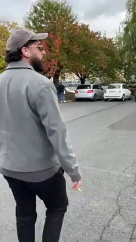 The video shows a man in a grey jacket, black jeans, and white sneakers, with a beard and sunglasses, standing in a parking lot on a cloudy day. He turns his head to look at the camera, smiles, and says something. There are cars parked in the background, along with trees that have autumn leaves. The man appears to be holding a cigarette in his hand.