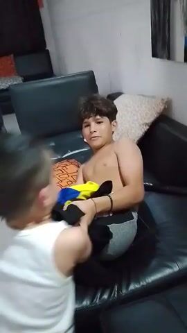 Two boys are playfully wrestling on a black leather couch indoors. The older boy, shirtless, is sitting while the younger one, wearing a white tank top and blue shorts, attempts to pin him down. Pillows with varied patterns are scattered on the couch. In the background, other furniture is visible, and the wall is painted white. The camera's focus shifts during the playful struggle.