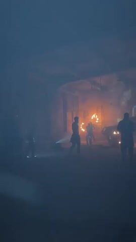 The video shows a dimly lit, smoky interior space with several people. In the center foreground, a person is silhouetted against a bright orange light source. To their right, another silhouetted person stands facing away from the camera. In the mid-ground, several other figures are visible, some interacting with what appear to be torches or fire. There are also objects that resemble vehicles or large equipment scattered throughout the space. The environment appears to be an industrial or abandoned building, with visible beams and brickwork in the background. The overall atmosphere is dark and dramatic, with a strong emphasis on the interplay of light and shadow.