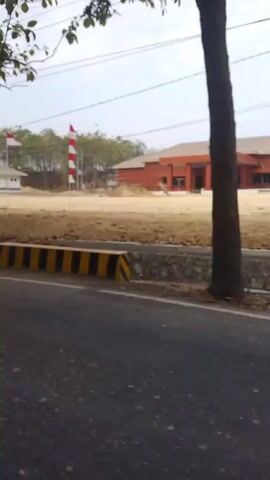The video captures a street-level view of a building and its surroundings. In the foreground, a section of an asphalt road is visible, with a yellow and black striped barrier running along its edge. Behind the barrier, there is a patch of dirt and vegetation, and a metal guardrail. Further back, several flags are flying, including the Indonesian flag. A large, brown building with a red roof is prominent in the middle ground, and appears to be under construction or recently built. More buildings, some with white walls and red roofs, are visible in the distance, along with trees and utility poles. The sky is overcast, suggesting a cloudy day. The camera appears to be mounted on a moving vehicle, as evidenced by the slight blur and the panning motion of the video.