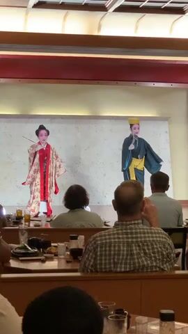 The video shows a performance by two individuals dressed in traditional East Asian attire on a stage. The person on the left wears a vibrant kimono with red and gold patterns and holds a fan. The person on the right wears a dark blue robe with a yellow sash and a yellow hat. They are both facing the audience, with the person on the left slightly turned to their left and the person on the right facing forward. The stage has a white backdrop with a textured surface. In the foreground, several audience members are seated at tables, with plates, glasses, and condiments visible. The setting appears to be a restaurant or dining area, with wooden tables and chairs. The lighting suggests an indoor environment, possibly with stage lighting illuminating the performers. The video captures a moment during the performance, with both individuals in various poses, suggesting a dance or theatrical presentation.