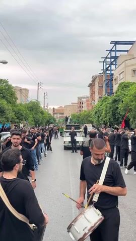 This is a vertical, eye-level, medium shot video of a religious procession on a street in Babol, Iran. The procession is taking place on a cloudy day, likely in the late afternoon or early evening, as indicated by the soft, diffused light and grey sky.

The primary subjects are a large group of men dressed in black clothing, participating in a procession. In the foreground, two men are visible, one on the left and one on the right, both holding drums. The man on the right has a white strap across his chest. The man on the left is holding a cymbal. The other men in the procession are also wearing black, some with long shirts and others with jackets. They appear to be marching or moving together in a coordinated manner.

The scene is a city street lined with buildings and trees. The buildings are multi-story structures with balconies and windows, typical of urban architecture. The street is paved and appears to be a public road, with some cars parked or moving in the background. There are power lines overhead, and streetlights are visible on the left side.

The activity taking place is clearly a procession, with the men moving down the street. The presence of drums and cymbals suggests a musical element to the event, likely a form of ceremonial drumming or chanting. The overall mood seems somber and respectful, given the all-black attire and the nature of religious processions. Some individuals in the crowd are looking towards the front of the procession, where a white car is visible with people on top, possibly a central figure or float. One person on the car appears to be speaking into a microphone. A red flag is visible on a pole in the distance.

There is no visible text in the video. The location cues, such as the architecture, street signs (though unreadable), and the presence of trees and vegetation, along with the information that the location is Babol, Iran, help to contextualize the scene as an urban religious event in that specific region.