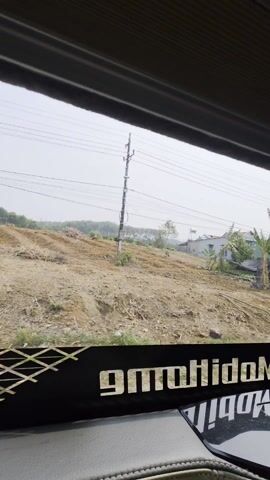 The video shows a view from inside a vehicle, likely a truck or van, as it travels along a road. The foreground includes a black dashboard and a decorative grill/screen bearing the word "mobihido" in reverse. The outside scenery visible through the windshield features rural landscapes with a mix of open dirt fields, houses, and various vegetation including banana trees, leafy trees, and other plants. Power lines are visible overhead. The sky is overcast, suggesting a cloudy or possibly hazy day. The camera appears relatively stable, capturing the passing scenery as the vehicle moves forward. There are no people visible in the frame.