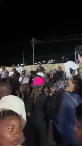 The video shows a crowd of people gathered outdoors at night, seemingly at a party or event. Many people are visible, with varying hairstyles and clothing. Several individuals are holding drinks, possibly beer bottles. One man is perched on a ledge or table while others interact around him. Overhead, there is a partial roof or awning structure adorned with colorful string lights. The atmosphere appears lively and social, with possible music or background noise contributing to the event's ambiance. The location seems to be urban, with building details visible against the night sky in the background.