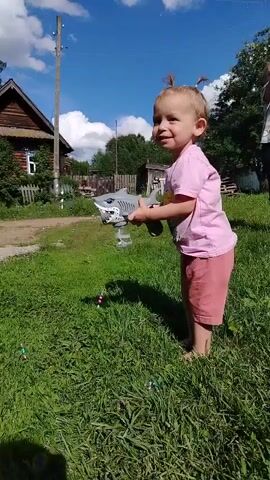 A young child, possibly a toddler, with blonde hair pulled into two small top knots, stands on a grassy lawn. The child is wearing a pink t-shirt and peach-colored shorts. They are holding a gray toy shark in their right hand, and in their left hand, they are holding what appears to be a bubble machine attached to the shark. The child is looking to the left of the frame and smiling. Bubbles are visible in the air around the child and the toy. In the background, there is a wooden house with a pitched roof and a white-trimmed window. Beyond the house, there are trees and a blue sky with white clouds. The scene appears to be outdoors on a sunny day.