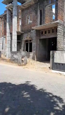 The video shows an unfinished brick building. The building is two stories high and has an open roof. The windows are bare openings, and there are no doors. The building is surrounded by a dirt and gravel area, and there is a paved road in front of it. A metal gate is visible on the right side of the building. The sky is blue with some clouds. The overall impression is one of a construction site that has been abandoned or is still in the early stages of development.