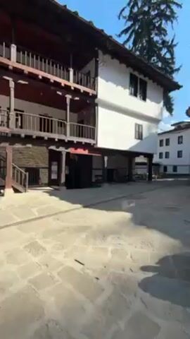 The video shows a courtyard with stone paving surrounded by buildings. A person is crouched, taking a photograph of a man standing in the courtyard. The camera pans and other people are seen walking across the courtyard. The day is sunny. The architectural style of the buildings appears historical.