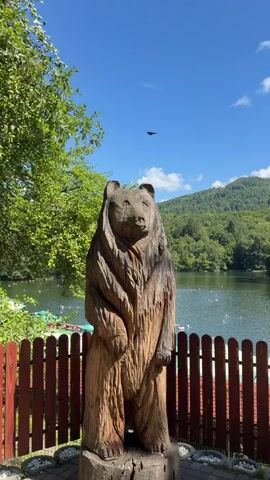 A full shot shows a wooden statue of a bear in the foreground. Beyond the statue, a lake is visible with people swimming and floating in it. The lake is surrounded by lush green trees and hills. Several green and white umbrellas are set up along the shore. The sky is a clear blue with a few scattered clouds.