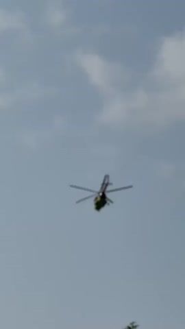 A helicopter is seen flying in the sky on a cloudy day. The helicopter is greenish-yellow with a red tail and is angled slightly downwards. The sky is a light blue with scattered white clouds. In the foreground, the top branches of a green tree are visible. The image appears to be taken from a low angle, looking up at the helicopter. The time of day appears to be daytime, likely afternoon, given the lighting. No people are visible in the frame. The setting is an outdoor environment, possibly a park or natural area, with trees. The activity taking place is simply the flight of the helicopter. No text is visible in the image.