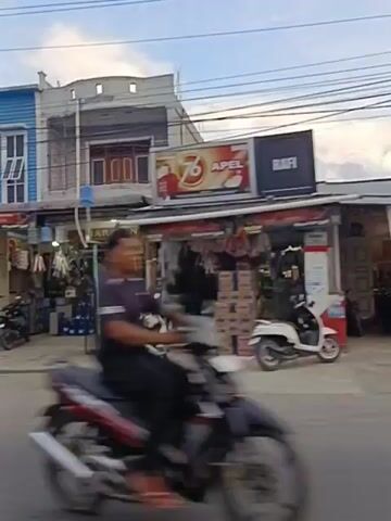 The video shows a street scene with several commercial establishments. On the left, there is a blue and white multi-story building with shutters. Next to it, a shop with an awning displays various items, including blue containers, a motorcycle, and what appears to be merchandise. Further down the street, there is a silver SUV parked. Another building features a red J&T Express sign and a gray shuttered shop. The next establishment has "Bee Cosmetics" signage. A small food stall is also visible on the left side of the frame. Several motorcycles are parked and one is being ridden. The environment appears to be an urban or suburban street with overhead wires and trees.
