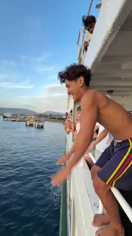 A young man jumps off the side of a boat into the water. He is wearing blue and yellow shorts and is shirtless. He enters the water headfirst, creating a large splash. The water is deep blue and there are other boats and land visible in the distance. The sky is a clear blue with some clouds. The video is shot from a high angle, looking down at the man as he jumps and then as he enters the water. The jump is captured in slow motion, allowing for a detailed view of the action. The overall impression is one of exhilaration and adventure.