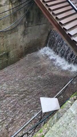 The media file shows a narrow, concrete channel with water flowing through it. The water is cascading over a small drop, creating a foamy white effect. Above the channel, there is a structure made of wooden planks, possibly a set of stairs or a walkway, angled downwards towards the water. Metal beams support this structure. The walls of the channel are rough, grey concrete. Several black cables run along the top and side of the channel. A small, rectangular white object, possibly a sign or a marker, is placed in the middle of the channel on the water's surface, held down by pipes. The water appears murky and a mix of grey and brown, with a rippling surface. The overall scene suggests an outdoor, possibly urban or semi-urban environment, given the presence of concrete and cables. The lighting suggests it could be daytime, but the weather is unclear. There is no discernible text visible in the media file. The presence of flowing water and the weathered appearance of the concrete and wood might suggest a location where water management or drainage is a factor, possibly in a hilly or mountainous region, consistent with the provided location context of Bhagsu, India, which is known for its waterfalls and natural scenery.