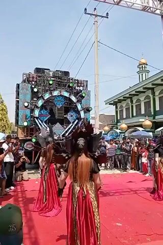 The video shows an outdoor performance with a large sound system setup. Several performers in elaborate costumes are on a red carpeted stage. In the foreground, three individuals are dressed in long, flowing red and gold gowns with dark, feathered headpieces. They appear to be dancing or performing. Behind them, a very large, multi-tiered sound system is visible, adorned with lights and a circular decorative element. To the left of the sound system, another performer is dressed in a white and silver costume that resembles a robot or creature, and is also performing. A crowd of spectators is gathered around the performance area, some holding cameras and phones. In the background, a mosque with a green dome and white walls is visible. The sky is clear and blue, suggesting it is daytime.