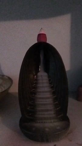 The video shows an object that appears to be a decorative piece, possibly an incense holder or diffuser. It is dark brown, egg-shaped, with a smaller, pink and white object placed on top. A pattern of concentric arches is visible within the object's structure, creating a tunnel-like illusion. The object is sitting on a white surface, likely a shelf or table, and is positioned against a white background. The lighting is somewhat dim.